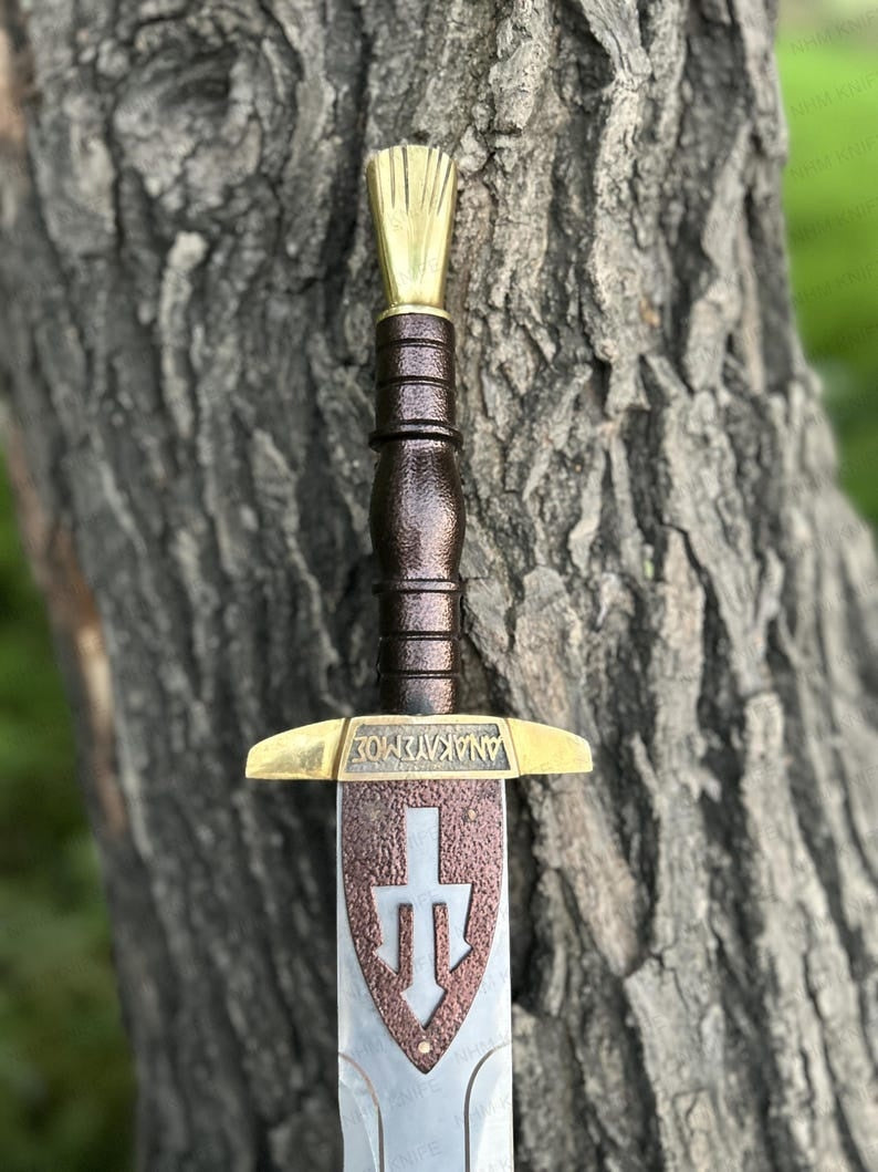 God of War Blade of Olympus replica sword leaning against a tree, showing the Greek inscriptions, bronze guard, and detailed steel blade.