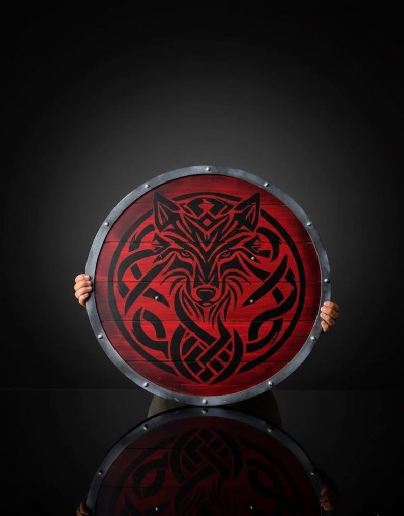"Handcrafted crimson red wooden Viking round shield featuring a black engraved Fenrir wolf face with Celtic knotwork and a riveted steel rim, held against a dark studio background."